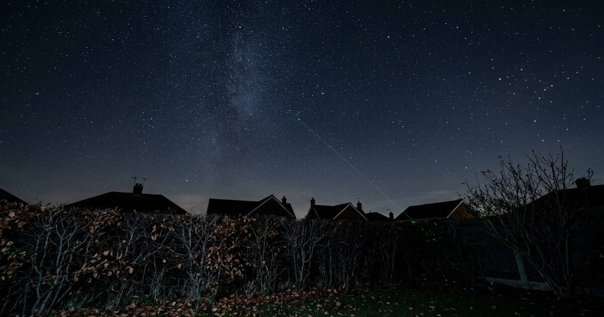 Groene laserstraal wijst vanuit een donkere achtertuin naar de sterrenhemel boven de daken
